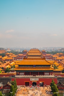 An iconic view of a vast, ancient architectural complex with large, ornate pavilions featuring distinctive yellow roofs, enclosed by a fortified wall. Crowds of visitors populate the main entrance area, hinting at the site's popularity as a cultural and historical attraction. The backdrop includes a hazy skyline under a clear blue sky.