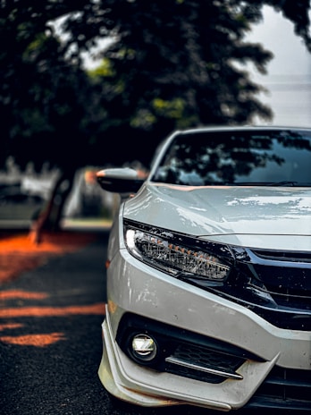 A close-up of a sleek white car positioned on a paved road. The image captures the front portion, focusing on the headlight and bumper. Surrounding the car are blurred trees, suggesting it's parked under their shade. The lighting creates a mix of sharp detail on the car and a soft bokeh effect in the background.