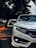 A close-up of a sleek white car positioned on a paved road. The image captures the front portion, focusing on the headlight and bumper. Surrounding the car are blurred trees, suggesting it's parked under their shade. The lighting creates a mix of sharp detail on the car and a soft bokeh effect in the background.