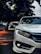 A close-up of a sleek white car positioned on a paved road. The image captures the front portion, focusing on the headlight and bumper. Surrounding the car are blurred trees, suggesting it's parked under their shade. The lighting creates a mix of sharp detail on the car and a soft bokeh effect in the background.