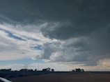 A funnel cloud protrudes from a large, dark storm cloud dominating the sky. The surrounding area consists of flat land with sparse trees and a few distant structures. The sky is a mix of dark and light clouds, with patches of blue and white visible in between.