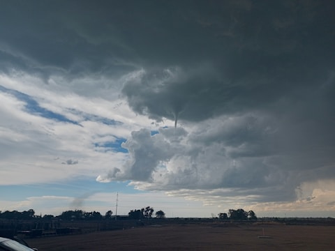 A funnel cloud protrudes from a large, dark storm cloud dominating the sky. The surrounding area consists of flat land with sparse trees and a few distant structures. The sky is a mix of dark and light clouds, with patches of blue and white visible in between.