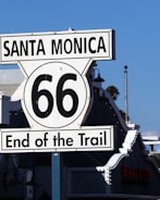 A Route 66 sign marks the end of the trail in Santa Monica. The sign is prominently displayed with a backdrop of blue sky and neighboring buildings. The iconic design features bold black text on a white background.