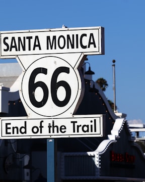 A Route 66 sign marks the end of the trail in Santa Monica. The sign is prominently displayed with a backdrop of blue sky and neighboring buildings. The iconic design features bold black text on a white background.