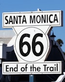 A road sign displaying 'Santa Monica' and '66 End of the Trail' against a clear blue sky. The iconic Route 66 symbol indicates the terminus of the historic highway.