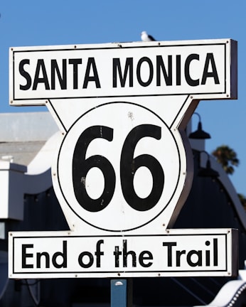 A road sign displaying 'Santa Monica' and '66 End of the Trail' against a clear blue sky. The iconic Route 66 symbol indicates the terminus of the historic highway.