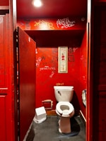 A dimly lit public restroom stall featuring red walls covered in various graffiti, stickers, and scribbles. In the center is a white toilet, and on the wall above it hangs a narrow framed image. A small trash bin is positioned beside the toilet. The overall scene is characterized by urban street art and a sense of disarray.