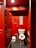 A dimly lit public restroom stall featuring red walls covered in various graffiti, stickers, and scribbles. In the center is a white toilet, and on the wall above it hangs a narrow framed image. A small trash bin is positioned beside the toilet. The overall scene is characterized by urban street art and a sense of disarray.