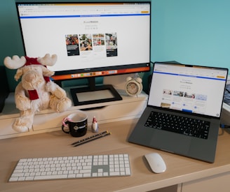 A workspace features a desktop with a large monitor and a laptop displaying a website. A plush toy moose wearing a winter hat and scarf sits beside the monitor. An alarm clock, a cup with a candy cane, and small decorative items are on the desk. A white keyboard and mouse are placed neatly on the wooden surface.