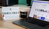 A desk with a lightbox displaying '#CHRISTMAS', a ceramic Santa figure, a black coffee mug with candy canes, and a laptop showing a webpage from Walls.io featuring various social media posts.