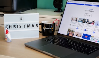 A desk with a lightbox displaying '#CHRISTMAS', a ceramic Santa figure, a black coffee mug with candy canes, and a laptop showing a webpage from Walls.io featuring various social media posts.