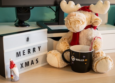 A plush reindeer wearing a Santa hat and scarf is sitting on a desk beside a lightbox displaying the words '# MERRY XMAS'. There is also a black mug with a company logo and a small figurine of a gnome with a red hat.