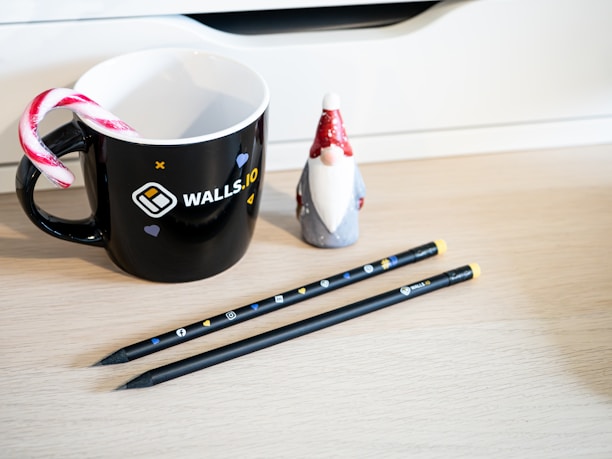A black mug featuring the logo 'WALLS.IO' with a candy cane placed inside, accompanied by a small figurine resembling a gnome. Two black pencils with social media icons are laid on the wooden surface.