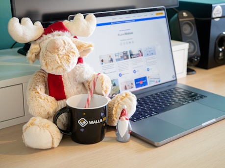 A plush toy resembling a reindeer or moose wearing a red and white Santa hat is sitting beside a laptop on a wooden desk. The laptop screen displays a web page. Next to the plush is a black mug with a candy cane inside, branded with 'Walls.io'. A small figurine of a gnome or Santa Claus is positioned next to the mug.