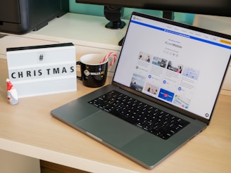 A modern desk setup featuring a laptop displaying a social media dashboard with the hashtag #LoveWallsio. To the left of the laptop is a black mug with a brand logo and two candy canes. A small Santa figurine and a lightbox sign with the word 'CHRISTMAS' and a hashtag are placed on the desk. The background includes a computer monitor and other office equipment.