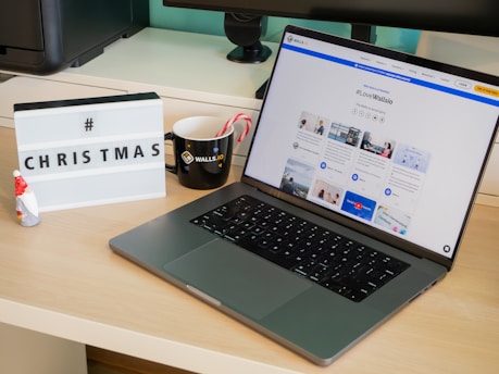 A modern desk setup featuring a laptop displaying a social media dashboard with the hashtag #LoveWallsio. To the left of the laptop is a black mug with a brand logo and two candy canes. A small Santa figurine and a lightbox sign with the word 'CHRISTMAS' and a hashtag are placed on the desk. The background includes a computer monitor and other office equipment.