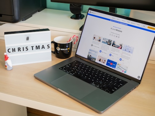 A modern desk setup featuring a laptop displaying a social media dashboard with the hashtag #LoveWallsio. To the left of the laptop is a black mug with a brand logo and two candy canes. A small Santa figurine and a lightbox sign with the word 'CHRISTMAS' and a hashtag are placed on the desk. The background includes a computer monitor and other office equipment.