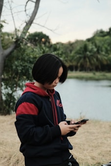 A person with short hair, dressed in a navy blue and red hoodie, is looking at a smartphone. The background features a tranquil outdoor setting with greenery and a body of water, suggesting a peaceful park or natural environment.