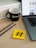 A workspace setup featuring a laptop with visible applications on the screen, a black mug with branding elements, two pencils, and a yellow sticky note displaying a hashtag symbol. These items are placed on a wooden surface with part of a white cabinet visible in the background.