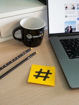 A workspace setup featuring a laptop with visible applications on the screen, a black mug with branding elements, two pencils, and a yellow sticky note displaying a hashtag symbol. These items are placed on a wooden surface with part of a white cabinet visible in the background.