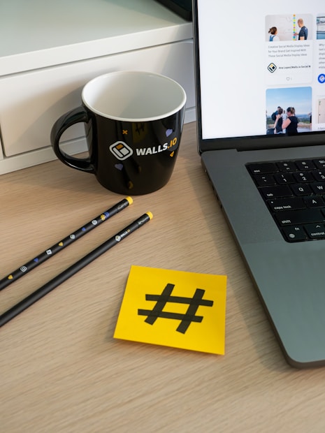 A workspace setup featuring a laptop with visible applications on the screen, a black mug with branding elements, two pencils, and a yellow sticky note displaying a hashtag symbol. These items are placed on a wooden surface with part of a white cabinet visible in the background.