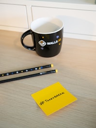 A black coffee mug with the logo and text 'WALLS.IO' sits on a light wooden desk. Nearby, two black pencils with yellow erasers and decorated with small icons are placed parallel to each other. A yellow sticky note with the handwritten hashtag '#TakeABreak' is on the table.