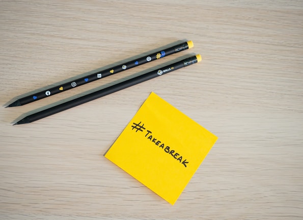Two black pencils with social media icons rest on a light wooden surface. Next to them is a yellow sticky note with the hashtag '#TAKEABREAK' written in black.