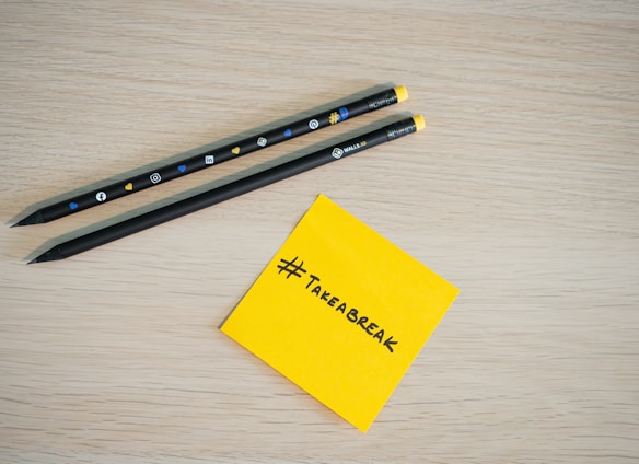 Two black pencils with social media icons rest on a light wooden surface. Next to them is a yellow sticky note with the hashtag '#TAKEABREAK' written in black.