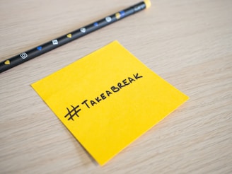 A yellow sticky note with the hashtag '#TakeABreak' written in black ink lies on a light wooden surface. Nearby, there is a black pencil featuring various social media icons and heart symbols.