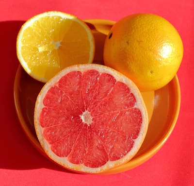 A close-up view of a halved grapefruit and orange positioned on a round, yellow plate. The vibrant red background enhances the bright colors of the citrus fruits.