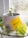 A small green plant grows in a textured, grayish pot located on a windowsill. Sunlight filters through the window, illuminating the vibrant leaves. A yellow sticky note with '#Growth' written on it is attached to the pot. The background shows the blurred outlines of a garden outside.