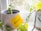 A small plant is potted in a grey ceramic planter placed on a windowsill. Another plant, with trailing vines and yellow-green leaves, is growing in a white pot next to it. A yellow sticky note with the word '#GROWTH' written on it is attached to the front of the grey planter. Outside the window, some blurry outdoor vegetation is visible.