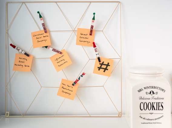 A wire wall grid holds several orange sticky notes attached with decorative wooden clips shaped like holiday figures. Each note has handwritten messages related to marketing campaigns such as 'Christmas Social Media Campaign' and 'New Year's Resolutions Campaign'. To the right of the grid, there is a large ceramic cookie jar labeled 'Mrs. Winterbottom's Delicious Traditions Cookies'. The setting appears clean and organized, suggesting a blend of festive planning and homey warmth.