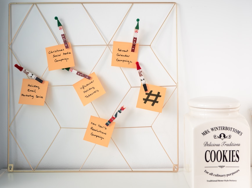 A wire wall grid holds several orange sticky notes attached with decorative wooden clips shaped like holiday figures. Each note has handwritten messages related to marketing campaigns such as 'Christmas Social Media Campaign' and 'New Year's Resolutions Campaign'. To the right of the grid, there is a large ceramic cookie jar labeled 'Mrs. Winterbottom's Delicious Traditions Cookies'. The setting appears clean and organized, suggesting a blend of festive planning and homey warmth.
