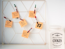 A wire wall grid holds several orange sticky notes attached with decorative wooden clips shaped like holiday figures. Each note has handwritten messages related to marketing campaigns such as 'Christmas Social Media Campaign' and 'New Year's Resolutions Campaign'. To the right of the grid, there is a large ceramic cookie jar labeled 'Mrs. Winterbottom's Delicious Traditions Cookies'. The setting appears clean and organized, suggesting a blend of festive planning and homey warmth.