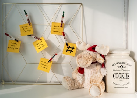 A decorative wire board displaying several yellow sticky notes, each with a handwritten campaign idea such as 'Christmas Social Media Campaign' and 'Advent Calendar Campaign'. Wooden clothespins with holiday-themed designs hold the notes. To the right of the board, a plush reindeer toy with a red scarf and hat leans against a large ceramic cookie jar labeled 'MRS. WINTERBOTTOM'S Delicious Traditions COOKIES'.