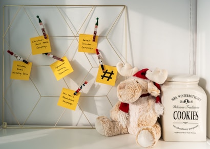 A decorative wire board displaying several yellow sticky notes, each with a handwritten campaign idea such as 'Christmas Social Media Campaign' and 'Advent Calendar Campaign'. Wooden clothespins with holiday-themed designs hold the notes. To the right of the board, a plush reindeer toy with a red scarf and hat leans against a large ceramic cookie jar labeled 'MRS. WINTERBOTTOM'S Delicious Traditions COOKIES'.