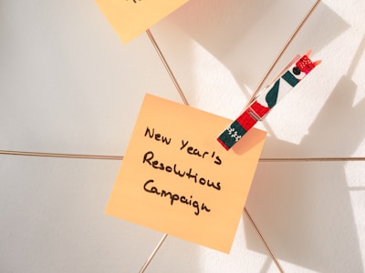 A note with the message 'New Year's Resolutions Campaign' is clipped onto a wire using a decorative clothespin. The clothespin has a festive pattern with colors such as red, green, and white. The background shows a light wall with shadows cast by natural light.