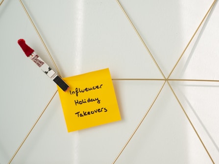 A yellow sticky note with the handwritten text 'Influencer Holiday Takeovers' is attached to a geometric wire grid. A decorative clothespin with a red and white pattern and a red pom-pom holds the note in place.