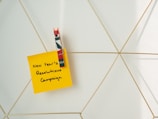 A yellow sticky note with the handwritten text 'New Year’s Resolutions Campaign' is attached to a geometric wire grid using a decorative clothespin with red, green, and white colors.
