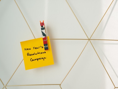 A yellow sticky note with the handwritten text 'New Year’s Resolutions Campaign' is attached to a geometric wire grid using a decorative clothespin with red, green, and white colors.