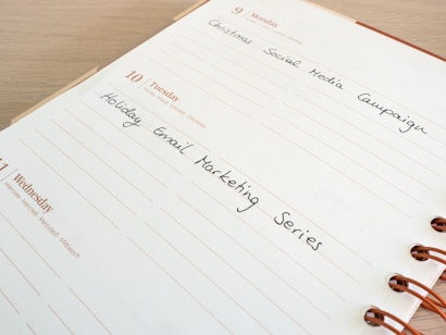 A close-up of a planner page, open to a week in December, with handwritten notes. The dates displayed include Monday, 9th December, and Tuesday, 10th December, with entries for 'Christmas Social Media Campaign' and 'Holiday Email Marketing Series'. The planner has a spiral binding and is set on a wooden surface.