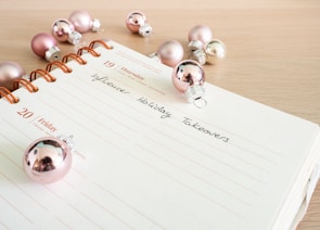 A planner or journal is open to a page showing Thursday the 19th and Friday the 20th. The pages are decorated with pink and silver Christmas ornaments scattered across a light wooden surface. The handwritten text on the page reads 'Influencer Holiday Takeovers'.