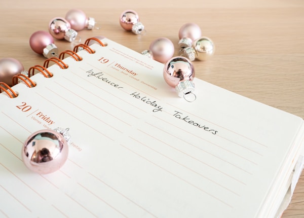 A planner or journal is open to a page showing Thursday the 19th and Friday the 20th. The pages are decorated with pink and silver Christmas ornaments scattered across a light wooden surface. The handwritten text on the page reads 'Influencer Holiday Takeovers'.