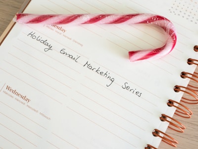 A candy cane rests on an open spiral notebook with an entry titled 'Holiday Email Marketing Series' written on it. The notebook page is white with lined sections, and the spiral binding is brown.