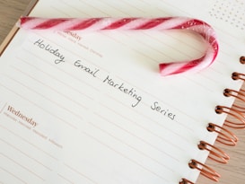 A candy cane rests on an open spiral notebook with an entry titled 'Holiday Email Marketing Series' written on it. The notebook page is white with lined sections, and the spiral binding is brown.