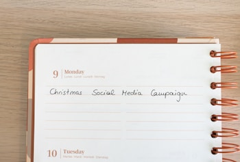 A calendar page for Monday, the 9th, featuring handwritten text that says 'Christmas Social Media Campaign.' The calendar is spiral-bound with orange rings, and the page has multilingual day headers.