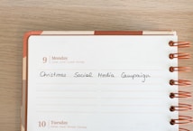 A calendar page for Monday, the 9th, featuring handwritten text that says 'Christmas Social Media Campaign.' The calendar is spiral-bound with orange rings, and the page has multilingual day headers.