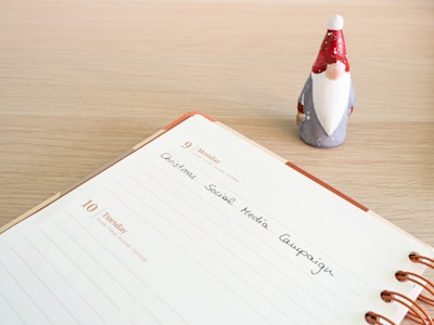 An open planner shows a page dated for Monday and Tuesday with the text 'Christmas Social Media Campaign' handwritten on it. A small figurine of a gnome with a red hat and white beard stands next to the planner on a wooden surface.