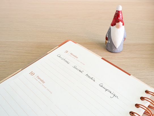 An open planner shows a page dated for Monday and Tuesday with the text 'Christmas Social Media Campaign' handwritten on it. A small figurine of a gnome with a red hat and white beard stands next to the planner on a wooden surface.